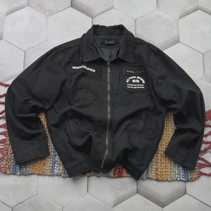May include: Black jacket with a zip fastening and patches with text. The jacket has a collar and two front pockets. The patches display the text "NARCOTRAFICO" and "69".