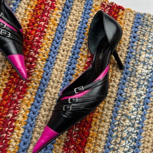 May include: A pair of black leather high-heeled shoes with pink accents and buckle details. The shoes are on a striped crocheted rug.
