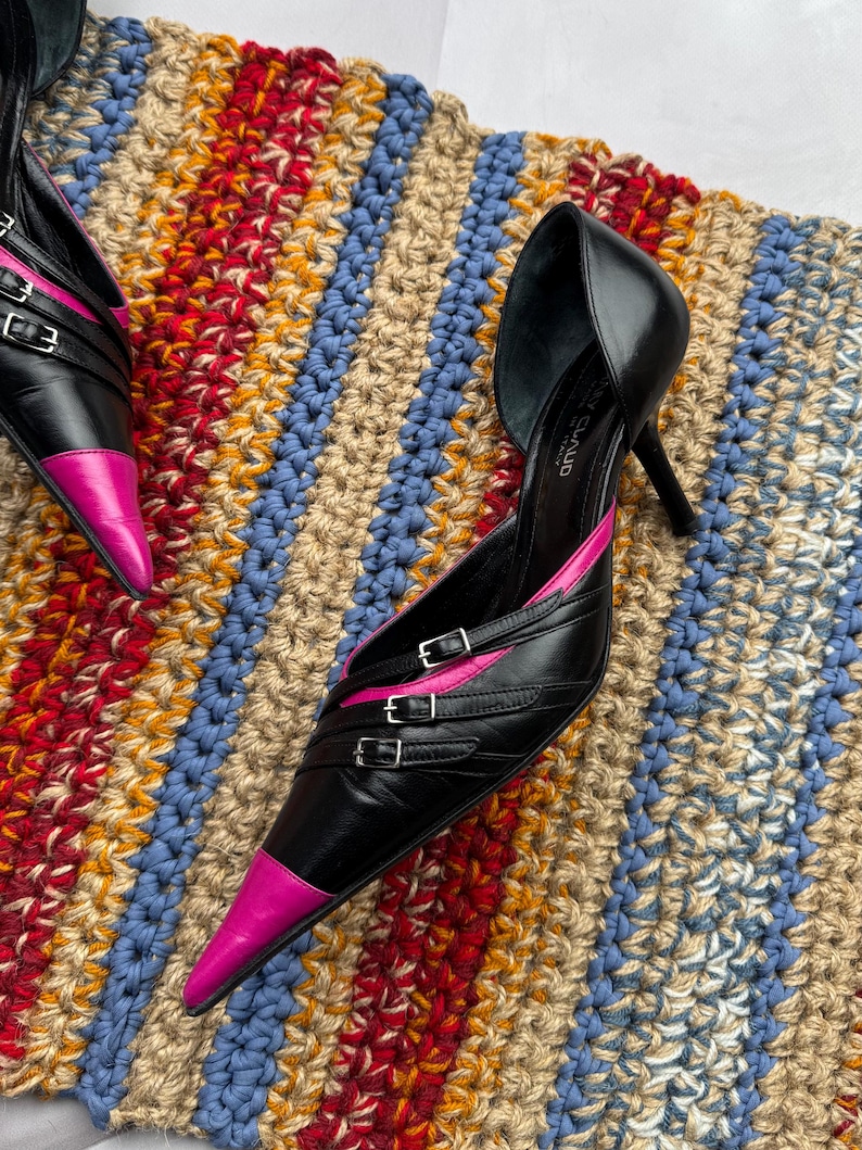 May include: A pair of black leather high-heeled shoes with pink accents and buckle details. The shoes are on a striped crocheted rug.