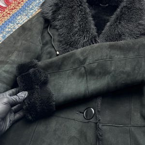 May include: A black shearling coat with a plush, furry collar and cuffs. The coat features a button closure and a soft, suede-like exterior. The interior lining is visible at the collar and cuffs, adding texture and warmth to the garment.