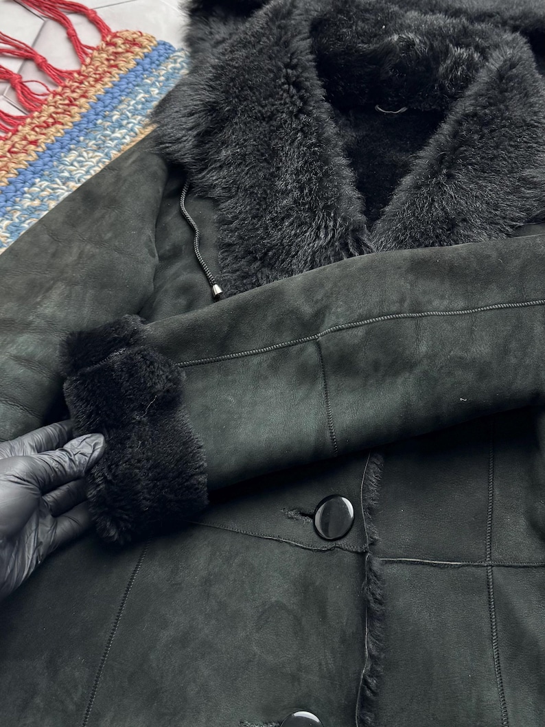 May include: A black shearling coat with a plush, furry collar and cuffs. The coat features a button closure and a soft, suede-like exterior. The interior lining is visible at the collar and cuffs, adding texture and warmth to the garment.