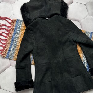 May include: A black suede coat with a hood trimmed with black fur. The coat has a button closure and fur trim on the cuffs. The coat is displayed on a colorful woven textile.