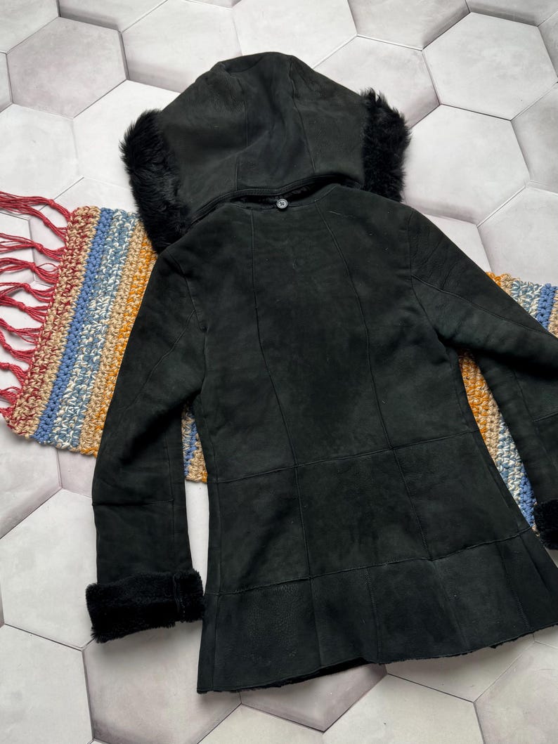 May include: A black suede coat with a hood trimmed with black fur. The coat has a button closure and fur trim on the cuffs. The coat is displayed on a colorful woven textile.