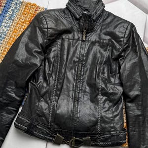 K&ouml;nnte beinhalten: Schwarze Lederjacke mit Rei&szlig;verschluss, dekorativen N&auml;hten und einem G&uuml;rtel in der Taille. Die Jacke hat einen Stehkragen mit gestickten Details. Ein stilvolles Oberbekleidungsst&uuml;ck.