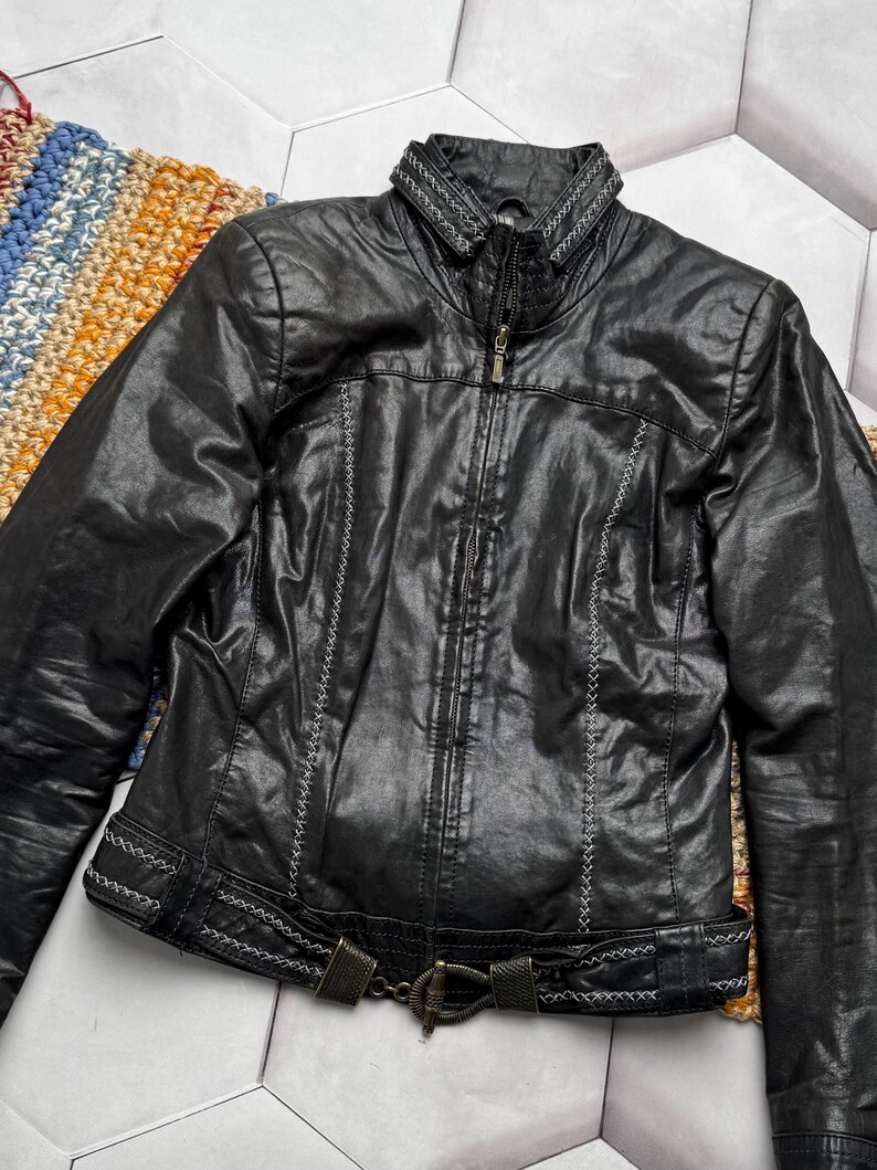 K&ouml;nnte beinhalten: Schwarze Lederjacke mit Rei&szlig;verschluss, dekorativen N&auml;hten und einem G&uuml;rtel in der Taille. Die Jacke hat einen Stehkragen mit gestickten Details. Ein stilvolles Oberbekleidungsst&uuml;ck.