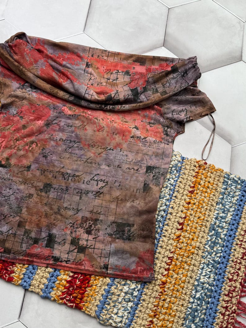 Op de afbeelding: Een top met gedrapeerde halslijn met een bloemen- en tekstprint in bruin-, rood- en paars tinten. Een veelkleurige gehaakte sjaal met franjes ligt naast de top. De top heeft een col en korte mouwen.