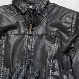 K&ouml;nnte beinhalten: Schwarze Lederjacke mit Stehkragen und Rei&szlig;verschluss. Die Jacke hat dekorative wei&szlig;e N&auml;hte entlang der N&auml;hte und des Kragens. Klassischer Stil.
