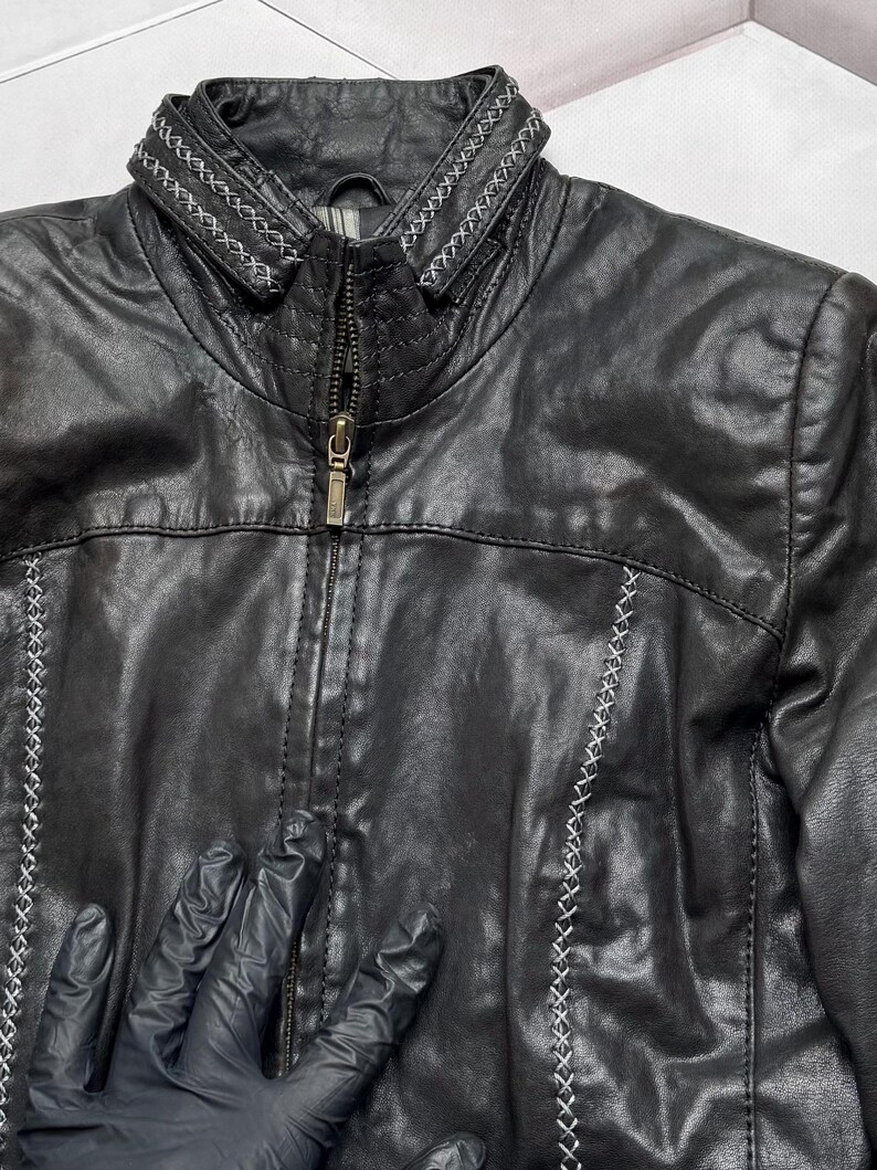 K&ouml;nnte beinhalten: Schwarze Lederjacke mit Stehkragen und Rei&szlig;verschluss. Die Jacke hat dekorative wei&szlig;e N&auml;hte entlang der N&auml;hte und des Kragens. Klassischer Stil.
