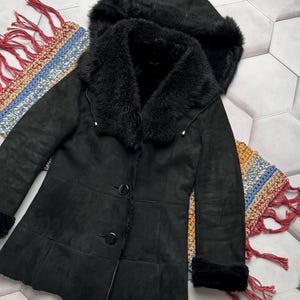 May include: Black hooded coat with a faux fur collar and trim. The coat features a button-up front and a tailored fit. The hood is also trimmed with faux fur. The coat is made of a soft, suede-like material.