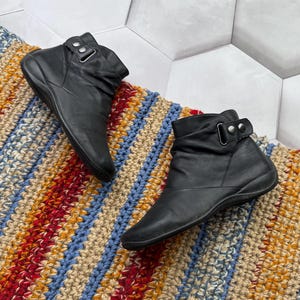 May include: A pair of black leather ankle boots with a buckle detail. The boots are displayed on a colorful, striped, crocheted rug. The rug features red, blue, yellow, and beige stripes. The boots have a low heel.