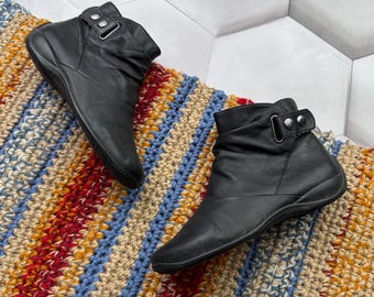 May include: A pair of black leather ankle boots with a buckle detail. The boots are displayed on a colorful, striped, crocheted rug. The rug features red, blue, yellow, and beige stripes. The boots have a low heel.