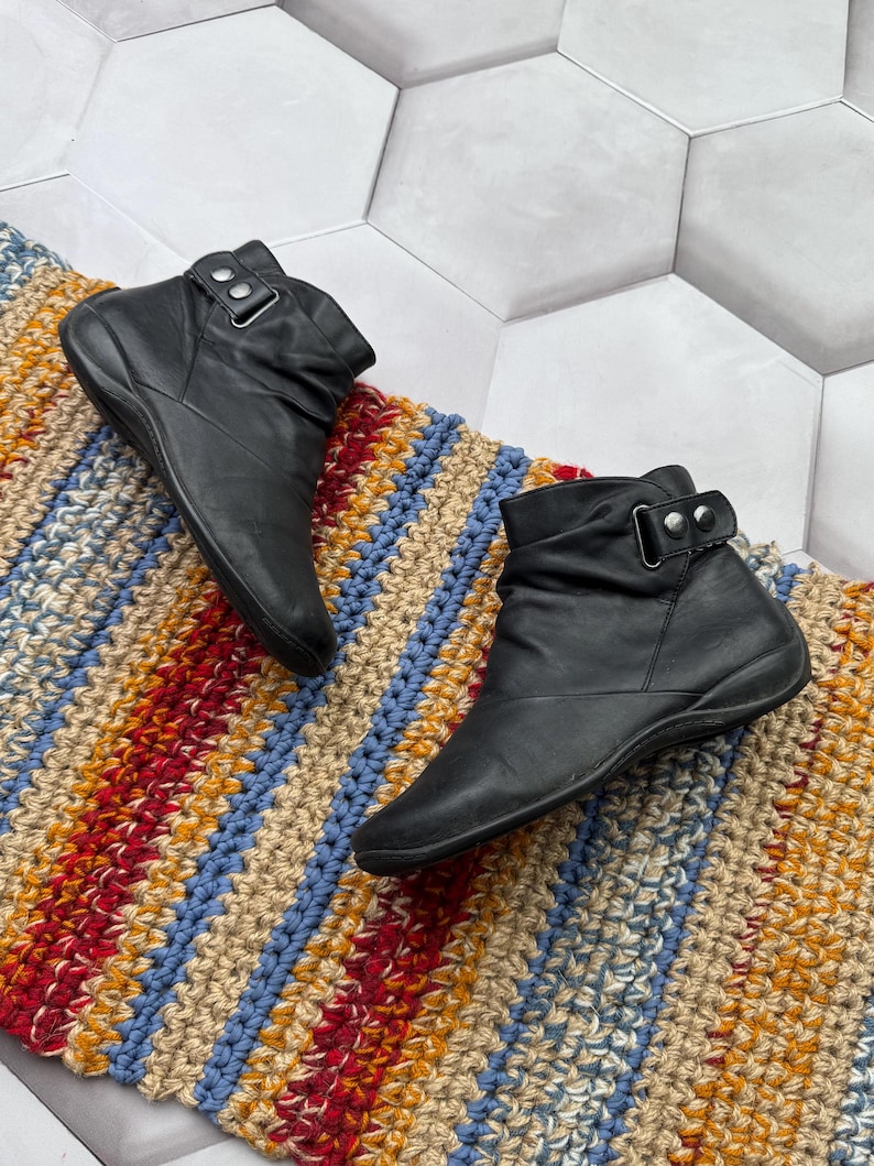 May include: A pair of black leather ankle boots with a buckle detail. The boots are displayed on a colorful, striped, crocheted rug. The rug features red, blue, yellow, and beige stripes. The boots have a low heel.