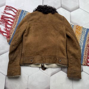 May include: A brown suede jacket with a black shearling collar and cream-colored lining. The jacket has a classic design with two front pockets and a zipper closure. The back of the jacket is visible, showcasing the tailored seams.