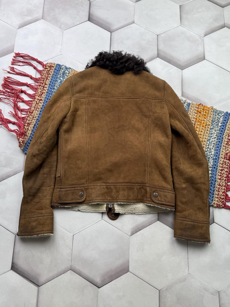 May include: A brown suede jacket with a black shearling collar and cream-colored lining. The jacket has a classic design with two front pockets and a zipper closure. The back of the jacket is visible, showcasing the tailored seams.
