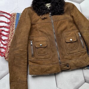 May include: Brown suede jacket with a black faux fur collar. The jacket features a zipper closure, two front pockets with button closures, and two side pockets with zippers. The jacket has a classic design.
