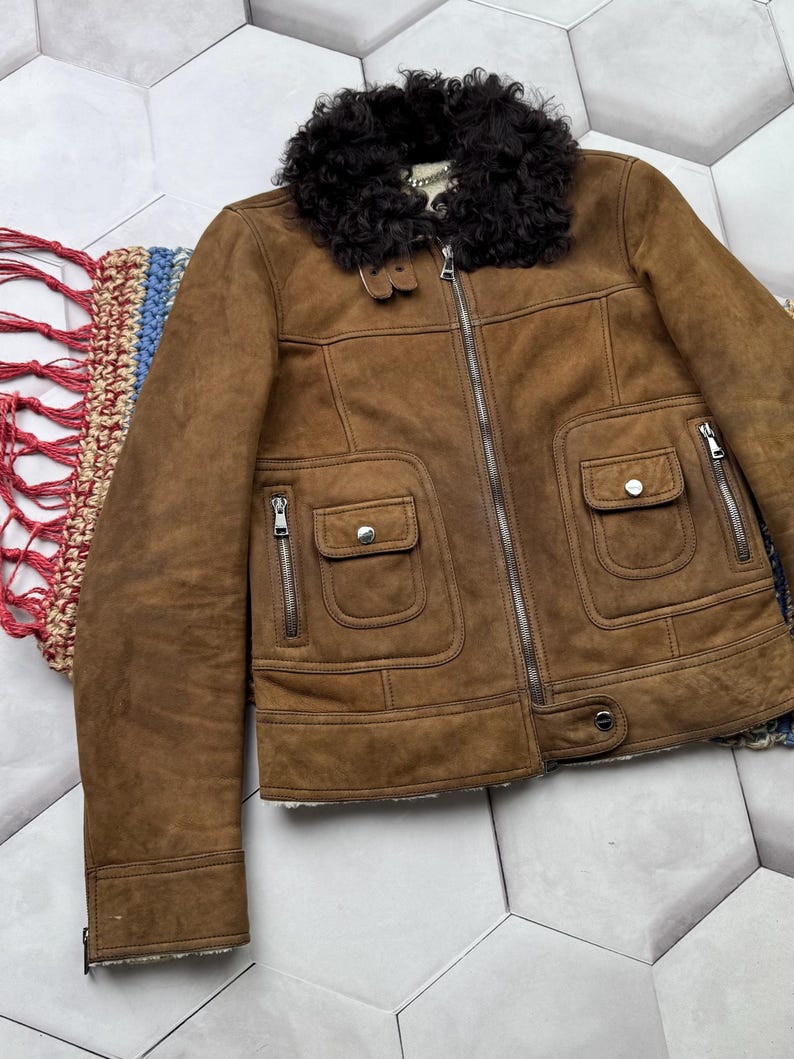 May include: Brown suede jacket with a black faux fur collar. The jacket features a zipper closure, two front pockets with button closures, and two side pockets with zippers. The jacket has a classic design.