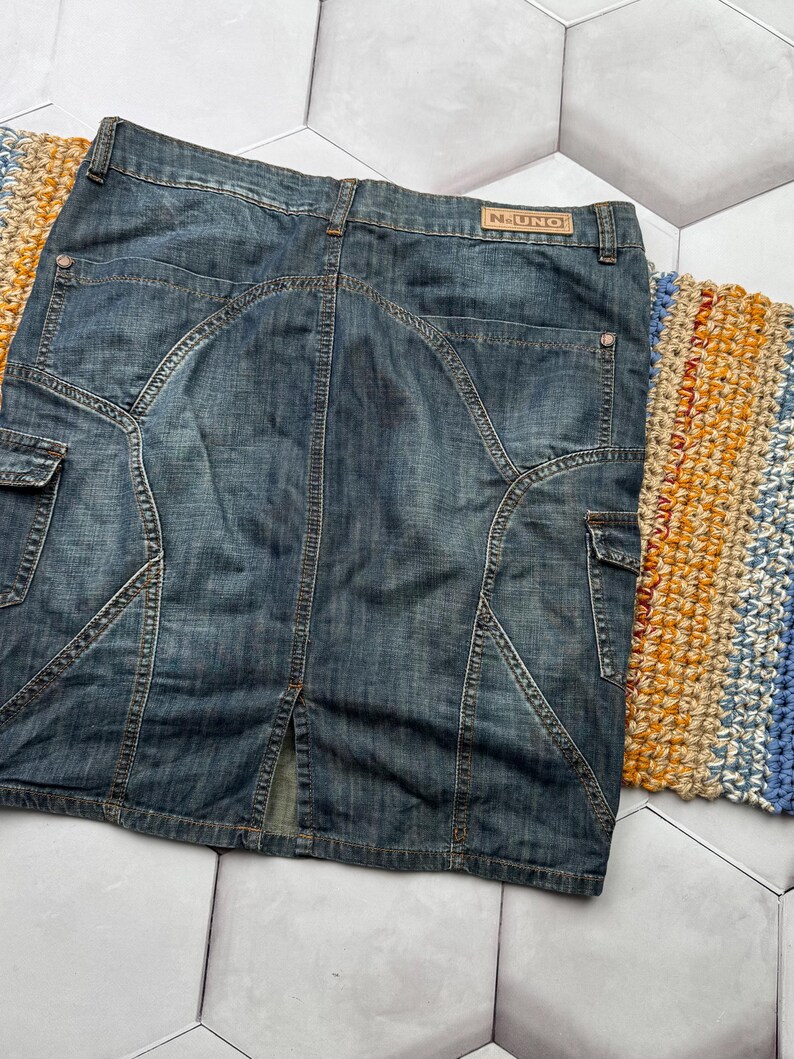 Puede incluir: Falda vaquera de color azul medio, de largo hasta la rodilla. Presenta bolsillos cargo, costuras decorativas y una peque&ntilde;a abertura en la parte trasera. La etiqueta de la marca "N-UNO" es visible en la cintura.