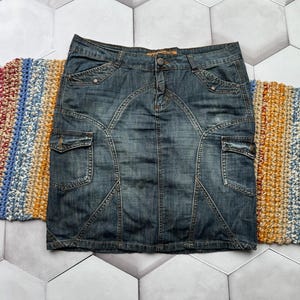 Puede incluir: Una falda vaquera en un lavado medio, con cierre de bot&oacute;n y dos bolsillos delanteros. La falda tiene un dise&ntilde;o ajustado con costuras decorativas y dos bolsillos de carga. La falda est&aacute; colocada sobre una superficie estampada.