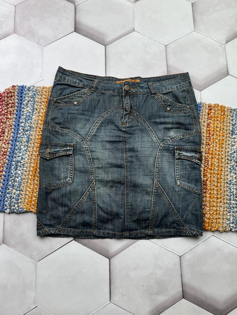 Puede incluir: Una falda vaquera en un lavado medio, con cierre de bot&oacute;n y dos bolsillos delanteros. La falda tiene un dise&ntilde;o ajustado con costuras decorativas y dos bolsillos de carga. La falda est&aacute; colocada sobre una superficie estampada.