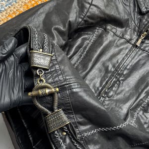 K&ouml;nnte beinhalten: Schwarze Kunstlederjacke mit dekorativen N&auml;hten und einem einzigartigen Verschluss. Die Jacke hat einen Rei&szlig;verschluss und detaillierte N&auml;hte entlang der N&auml;hte. Der Verschluss ist ein verziertes Metalldesign. Ein stilvolles Oberbekleidungsst&uuml;ck.