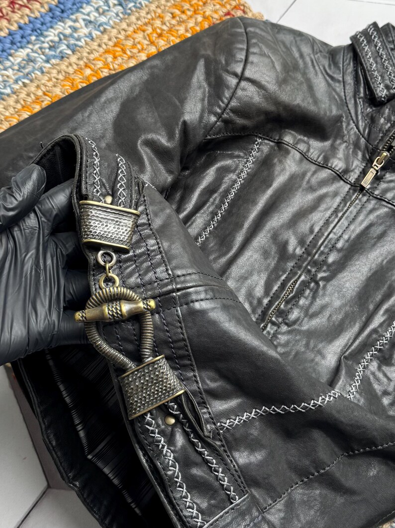 K&ouml;nnte beinhalten: Schwarze Kunstlederjacke mit dekorativen N&auml;hten und einem einzigartigen Verschluss. Die Jacke hat einen Rei&szlig;verschluss und detaillierte N&auml;hte entlang der N&auml;hte. Der Verschluss ist ein verziertes Metalldesign. Ein stilvolles Oberbekleidungsst&uuml;ck.