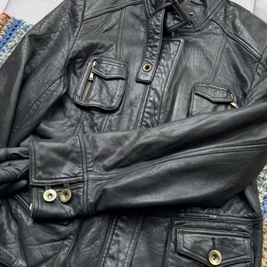 May include: A black leather jacket with a zipper closure and multiple pockets. The jacket has a stand-up collar and gold-toned hardware.