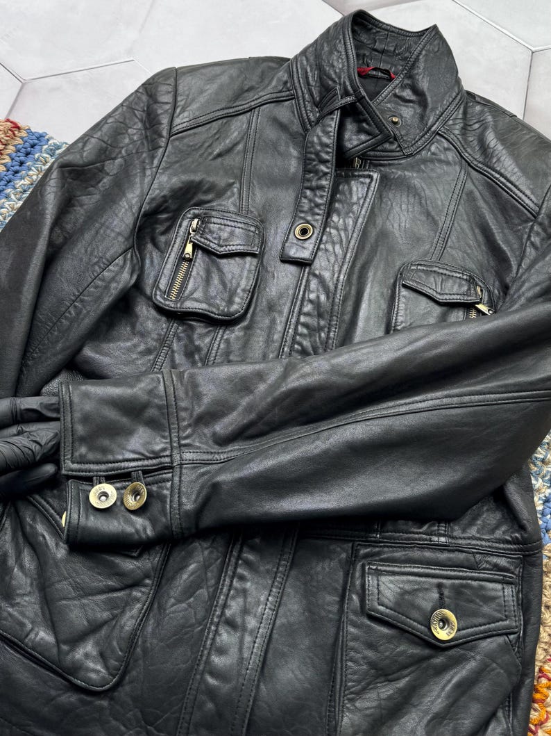 May include: A black leather jacket with a zipper closure and multiple pockets. The jacket has a stand-up collar and gold-toned hardware.