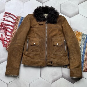 May include: A brown suede jacket with a black fur collar. The jacket has a zipper closure, two front pockets with zippers, and a button detail at the waist. The jacket is displayed on a patterned rug.