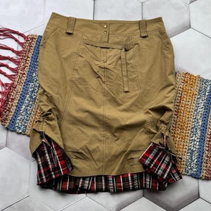 May include: A tan skirt with a plaid hem and cuffs. The skirt has a fitted silhouette, a belt, and pockets. The cuffs are red, black, and white plaid. The skirt is displayed on a gray and white tiled surface.