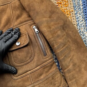 May include: A brown suede jacket with a zipper closure and two front pockets. The jacket has a classic design with a soft, textured appearance. The sleeve is visible, and the jacket appears to be well-made.