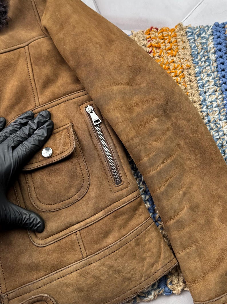 May include: A brown suede jacket with a zipper closure and two front pockets. The jacket has a classic design with a soft, textured appearance. The sleeve is visible, and the jacket appears to be well-made.