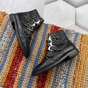 May include: A pair of black leather ankle boots with multiple silver buckles and studded details. The boots are displayed on a colorful, striped rug with red, blue, and tan hues.