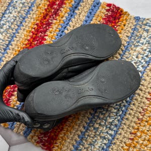 May include: Black leather shoes with the brand name "Clarks" visible on the soles. The soles have a textured design. The shoes are on a colorful, striped rug.