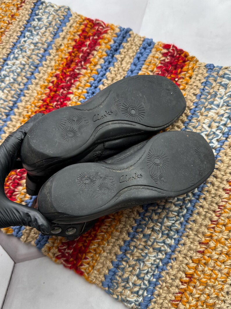 May include: Black leather shoes with the brand name "Clarks" visible on the soles. The soles have a textured design. The shoes are on a colorful, striped rug.