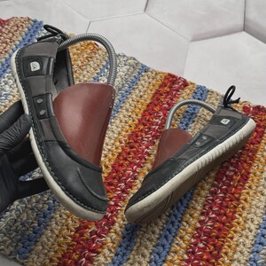 May include: A pair of dark gray leather shoes featuring a low-profile design, white rubber outsoles, and a dark red plastic shoe tree insert. The shoes have a small silver logo emblem on the side and a black pull tab at the heel for easy wear.
