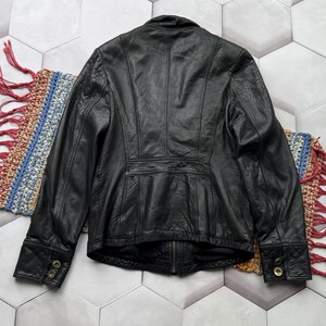 May include: A black leather jacket with a zipper closure and button accents on the cuffs. The jacket has a fitted silhouette and a stand-up collar.