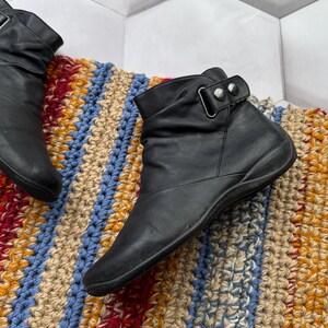 May include: Black leather ankle boots with a strap and buckle detail. The boots have a low heel and a rounded toe. The boots are on a colorful, textured rug with blue, red, yellow, and beige stripes.
