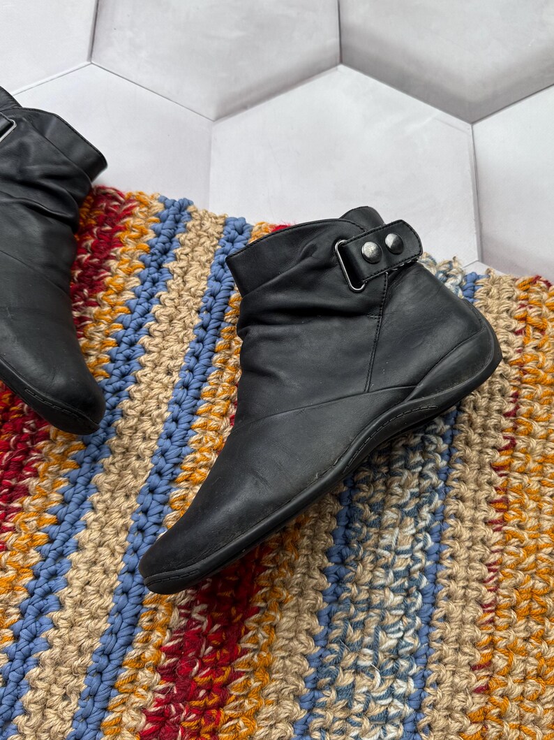 May include: Black leather ankle boots with a strap and buckle detail. The boots have a low heel and a rounded toe. The boots are on a colorful, textured rug with blue, red, yellow, and beige stripes.