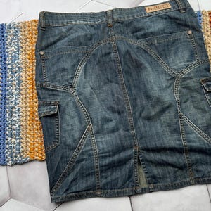 Puede incluir: Falda vaquera de lavado medio con dise&ntilde;o hasta la rodilla. Cuenta con bolsillos cargo, abertura trasera y costuras decorativas. La etiqueta "N-UNO" es visible en la cintura. La falda est&aacute; colocada sobre una superficie estampada.