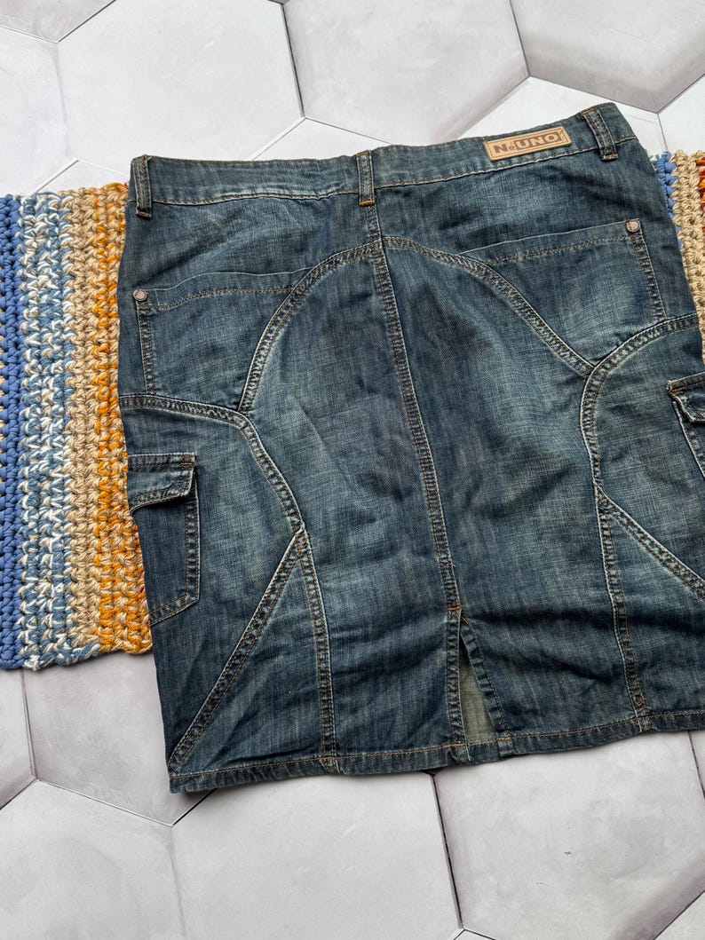 Puede incluir: Falda vaquera de lavado medio con dise&ntilde;o hasta la rodilla. Cuenta con bolsillos cargo, abertura trasera y costuras decorativas. La etiqueta "N-UNO" es visible en la cintura. La falda est&aacute; colocada sobre una superficie estampada.