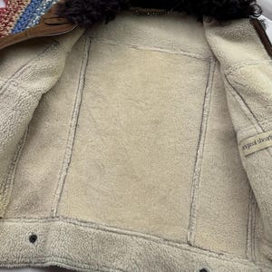 May include: A brown shearling jacket with a black fur collar. The interior is lined with cream-colored shearling. The jacket has a zipper closure and two buttons at the bottom. The label reads "original shearling".