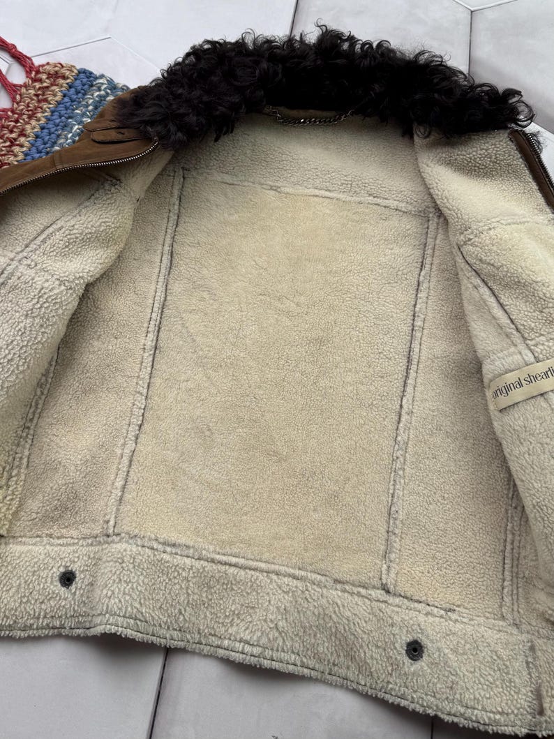 May include: A brown shearling jacket with a black fur collar. The interior is lined with cream-colored shearling. The jacket has a zipper closure and two buttons at the bottom. The label reads "original shearling".