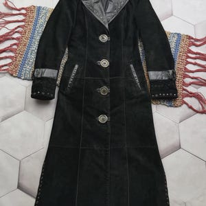 Può includere: Un cappotto lungo in camoscio nero con colletto a revers e dettagli con occhielli in metallo argentato. Il cappotto presenta quattro grandi bottoni frontali, maniche lunghe con polsini con fibbia e pannelli verticali fino all'orlo con occhielli.