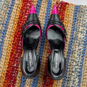 May include: A pair of black leather high-heeled shoes with pink pointed toes and double buckles. The shoes are on a striped rug and say "Mary Claud Made in Italy" on the inside.