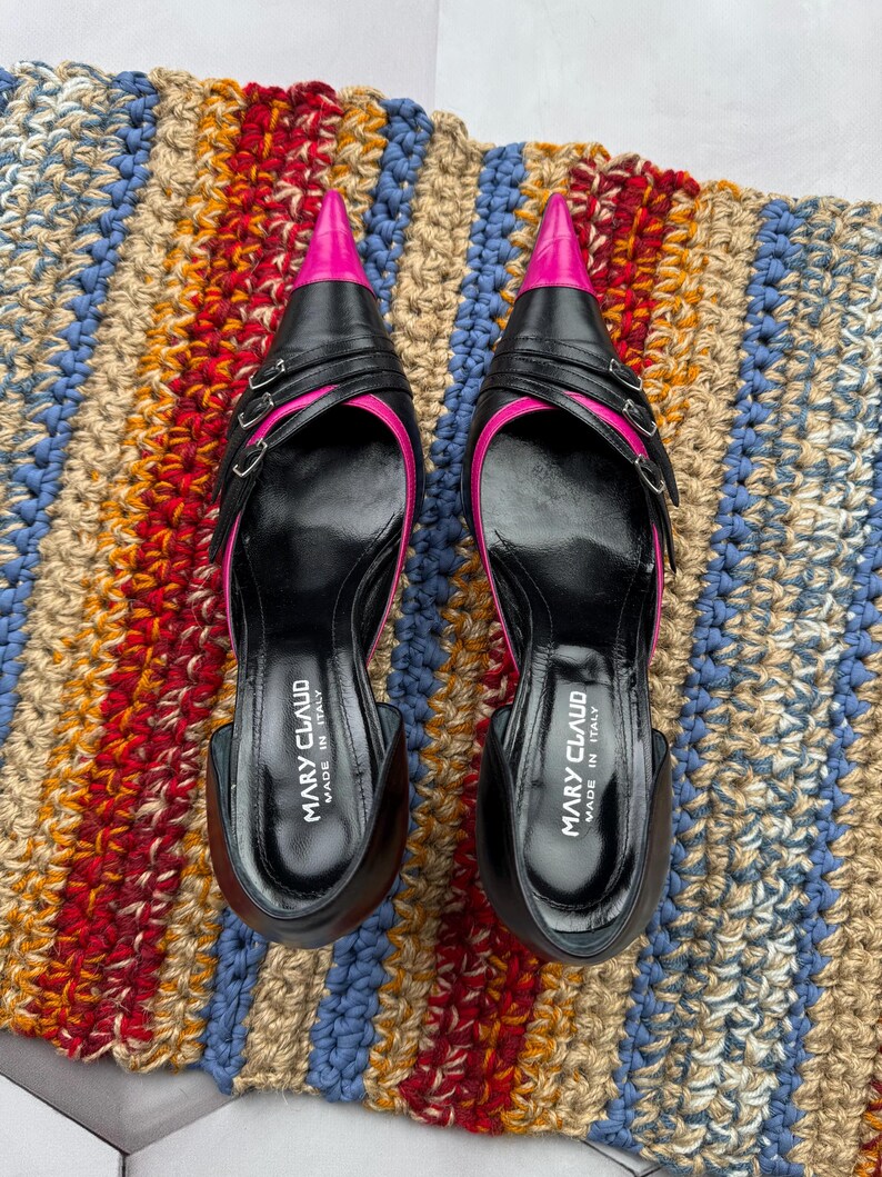 May include: A pair of black leather high-heeled shoes with pink pointed toes and double buckles. The shoes are on a striped rug and say "Mary Claud Made in Italy" on the inside.