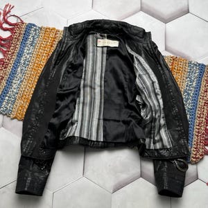 K&ouml;nnte beinhalten: Schwarze Lederjacke mit gestreiftem Futter. Die Jacke hat ein klassisches Design mit Kragen, Rei&szlig;verschluss und Schnallendetails an den &Auml;rmeln. Das Innenfutter weist ein schwarz-wei&szlig;-graues Streifenmuster auf.