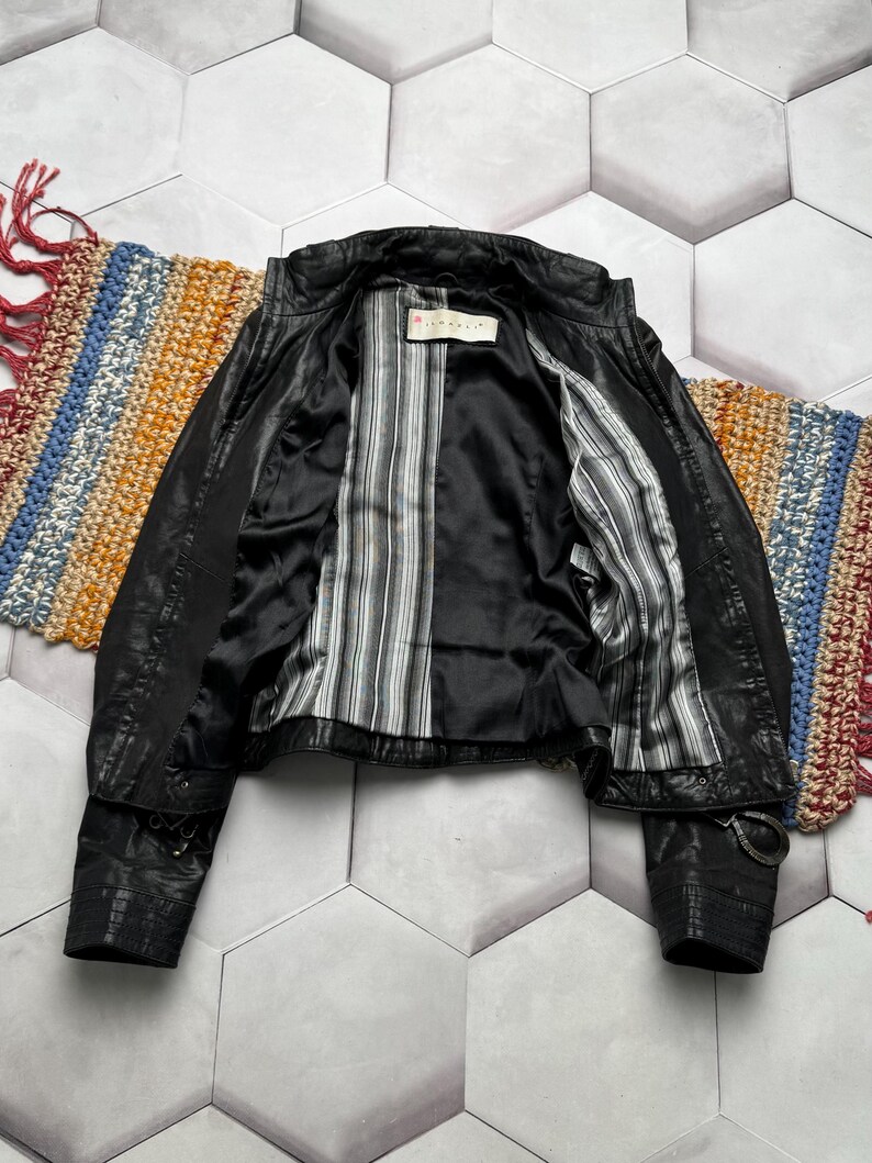 K&ouml;nnte beinhalten: Schwarze Lederjacke mit gestreiftem Futter. Die Jacke hat ein klassisches Design mit Kragen, Rei&szlig;verschluss und Schnallendetails an den &Auml;rmeln. Das Innenfutter weist ein schwarz-wei&szlig;-graues Streifenmuster auf.