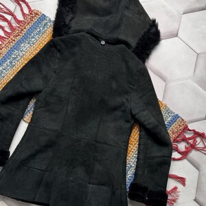 May include: A black suede jacket with a hood trimmed with black fur. The jacket has a button closure and a flared hem. The sleeves have fur cuffs. The jacket is displayed on a colorful, patterned textile.