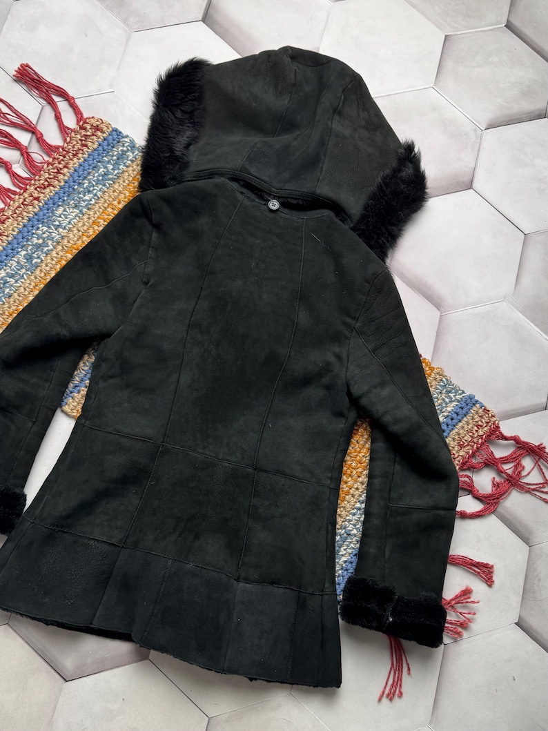 May include: A black suede jacket with a hood trimmed with black fur. The jacket has a button closure and a flared hem. The sleeves have fur cuffs. The jacket is displayed on a colorful, patterned textile.