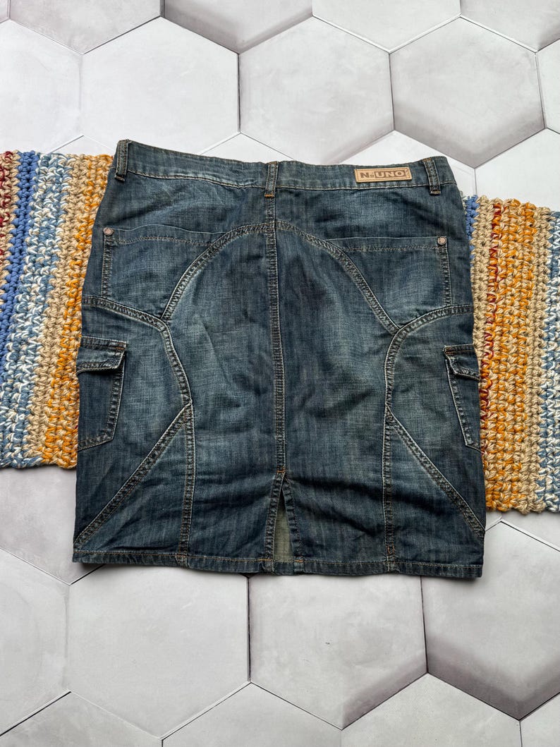 Puede incluir: Falda vaquera azul descolorido. Cuenta con bolsillos cargo, costuras decorativas y una abertura trasera. La etiqueta "N-UNO" es visible en la cintura. La falda es de estilo hasta la rodilla.